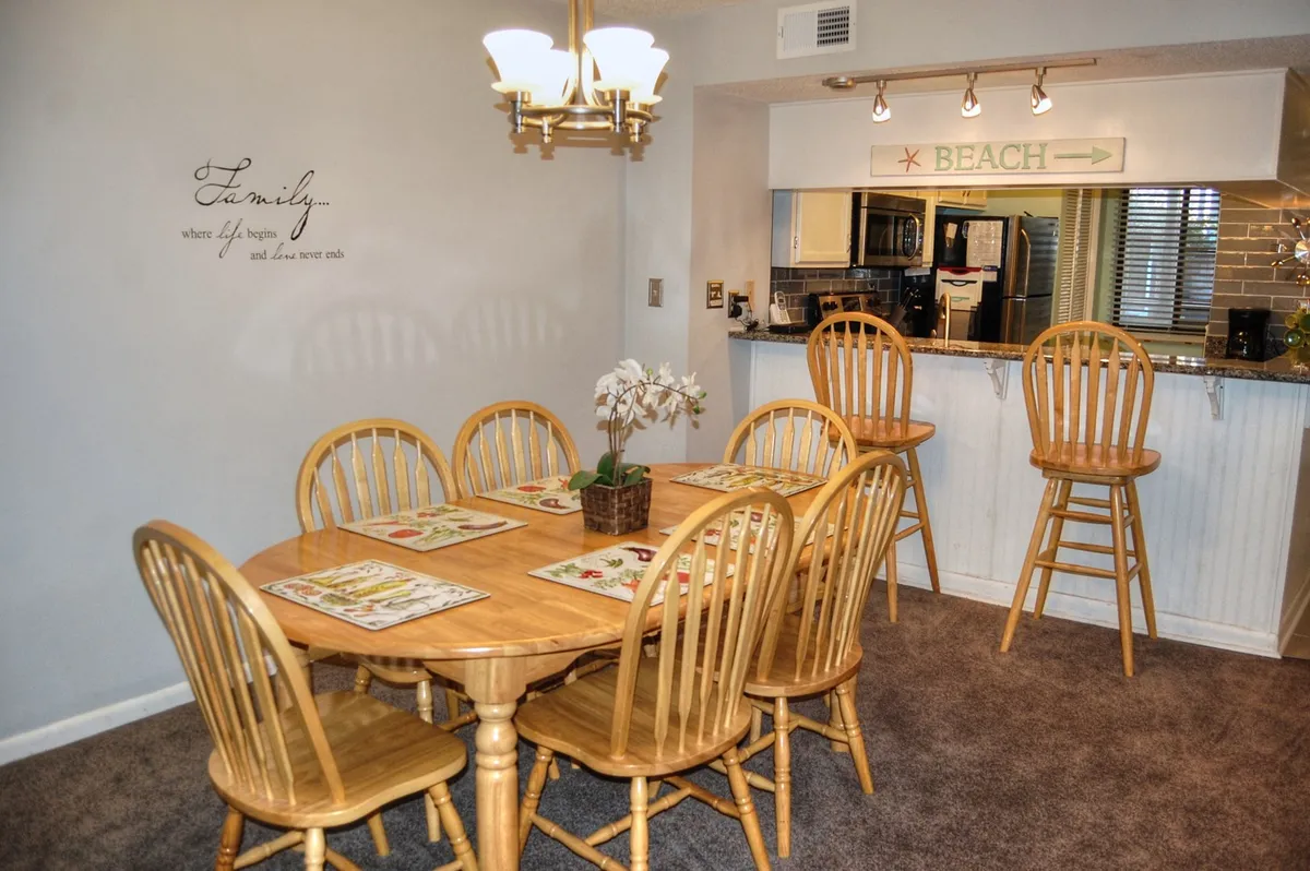 A stylish dining area with a family quote on the wall, elegant chandelier, and a wooden dining table set for meals or games. A kitchen bar and decorative plant complete the scene, emphasizing the space's warmth and versatility.