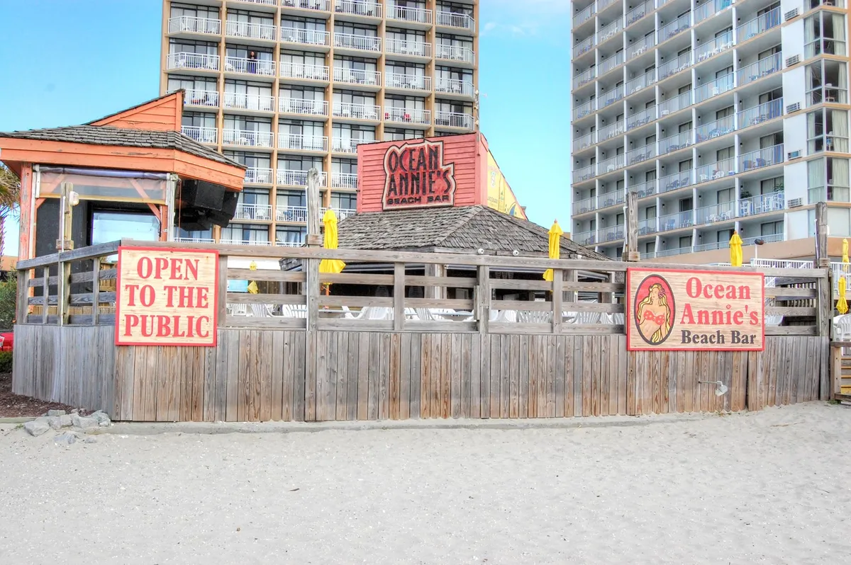 Ocean Annie's Beach Bar is highlighted with its welcoming signage, yellow umbrellas, and open-air setup ideal for socializing, dining outdoors, and enjoying live music or the sound of the waves. The mood is relaxed and festive.