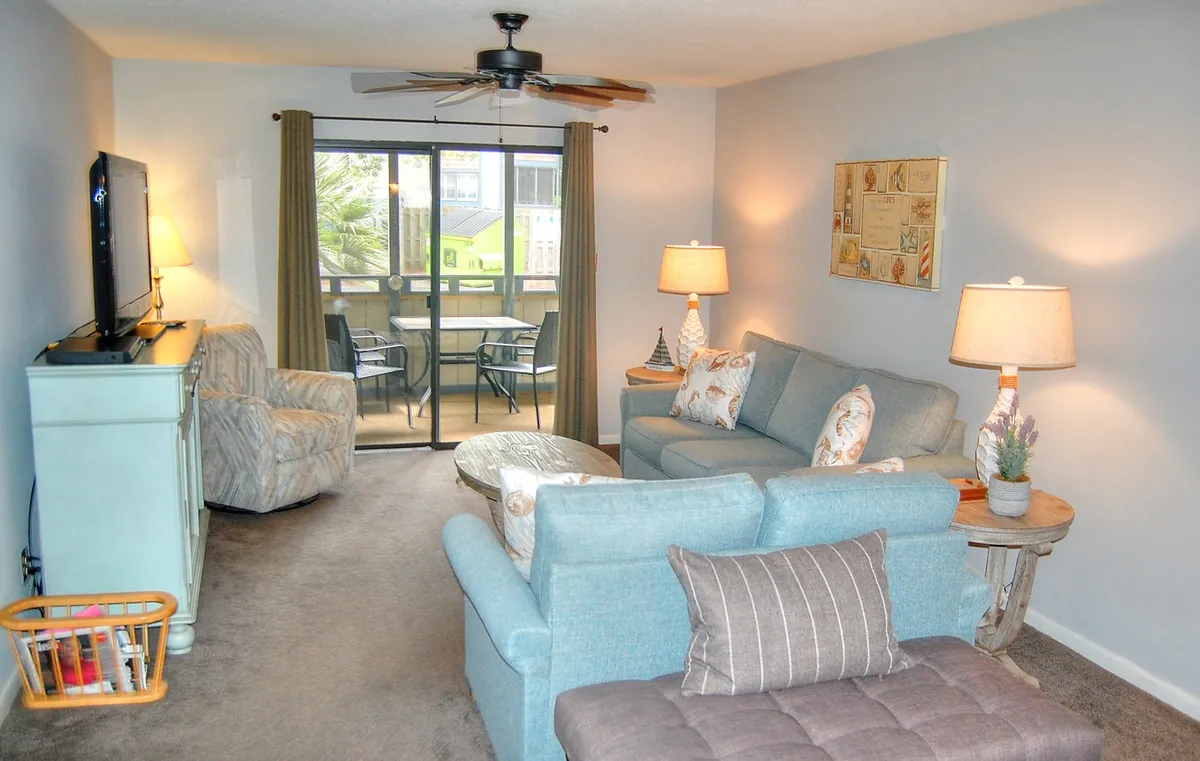 A sunlit living room featuring a sofa, armchair, coffee table, and TV, with a sliding glass door bathing the space in natural light. Warm lighting and a relaxed arrangement set the stage for both lively conversations and quiet relaxation.