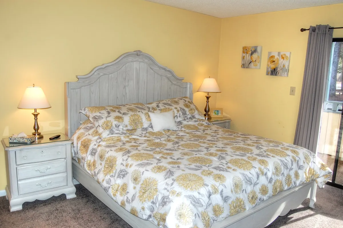 A cozy bedroom featuring a bed with floral bedding, flanked by nightstands with matching lamps and accent artwork above. Natural light streams through the window, enhancing the inviting atmosphere.