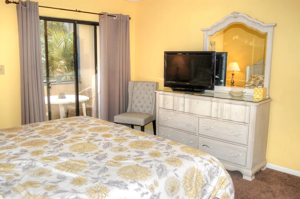 A tranquil bedroom with a floral-patterned bed, a dressing table and mirror, a television, and a large window with curtains. The space exudes comfort and privacy.