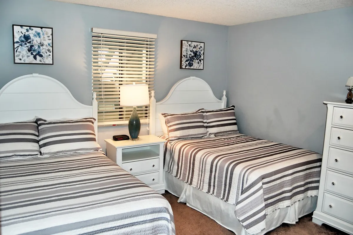 A bright, comfortable bedroom with two beds featuring striped bedding, a shared nightstand with a lamp, and decorative wall art. The room feels airy and designed for shared occupancy.