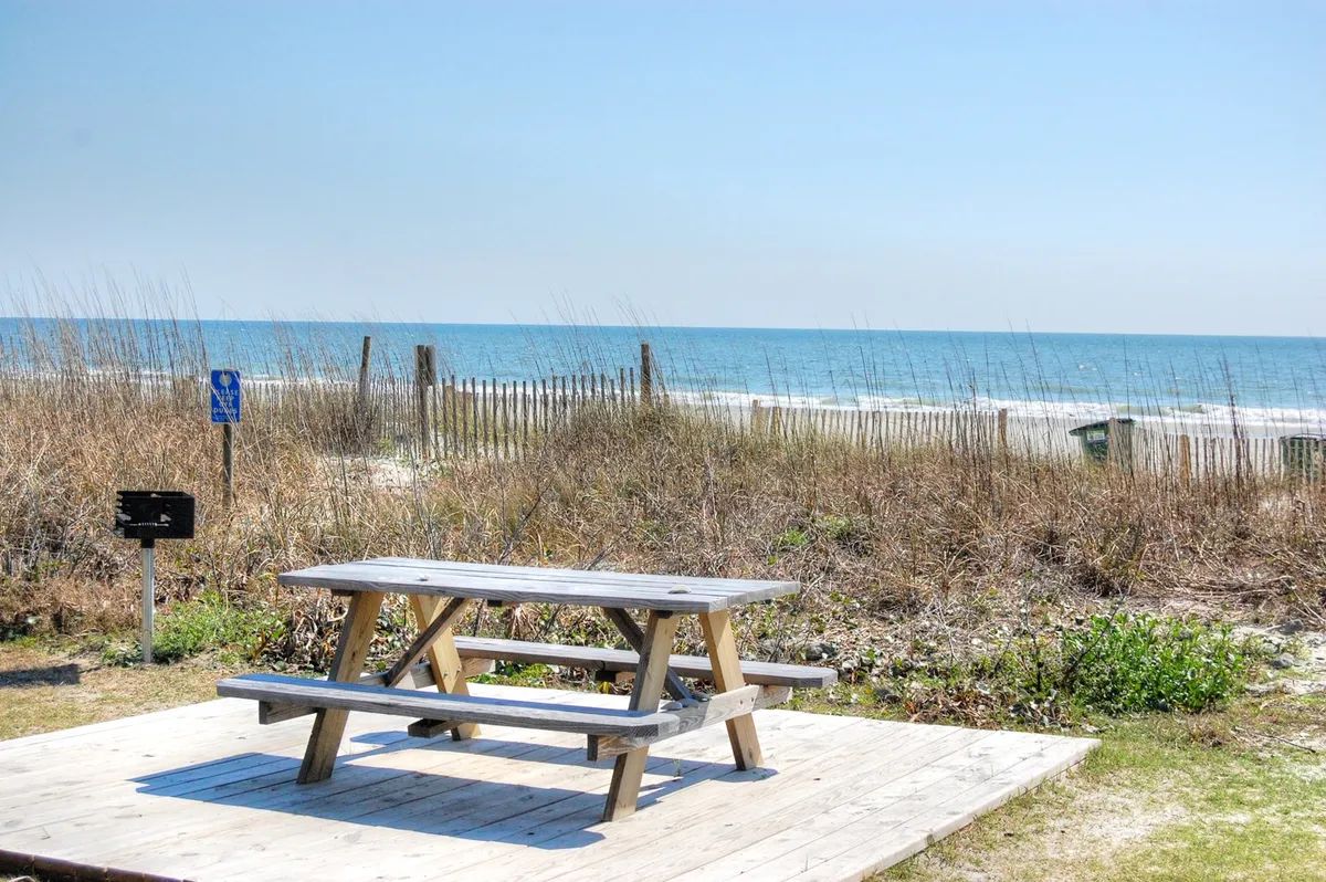 A peaceful picnic table overlooks the ocean, framed by dune grass and a wooden fence. The view suggests easy access to the quiet beachfront, with space for family picnics, impromptu games, and relaxation just steps from the water.