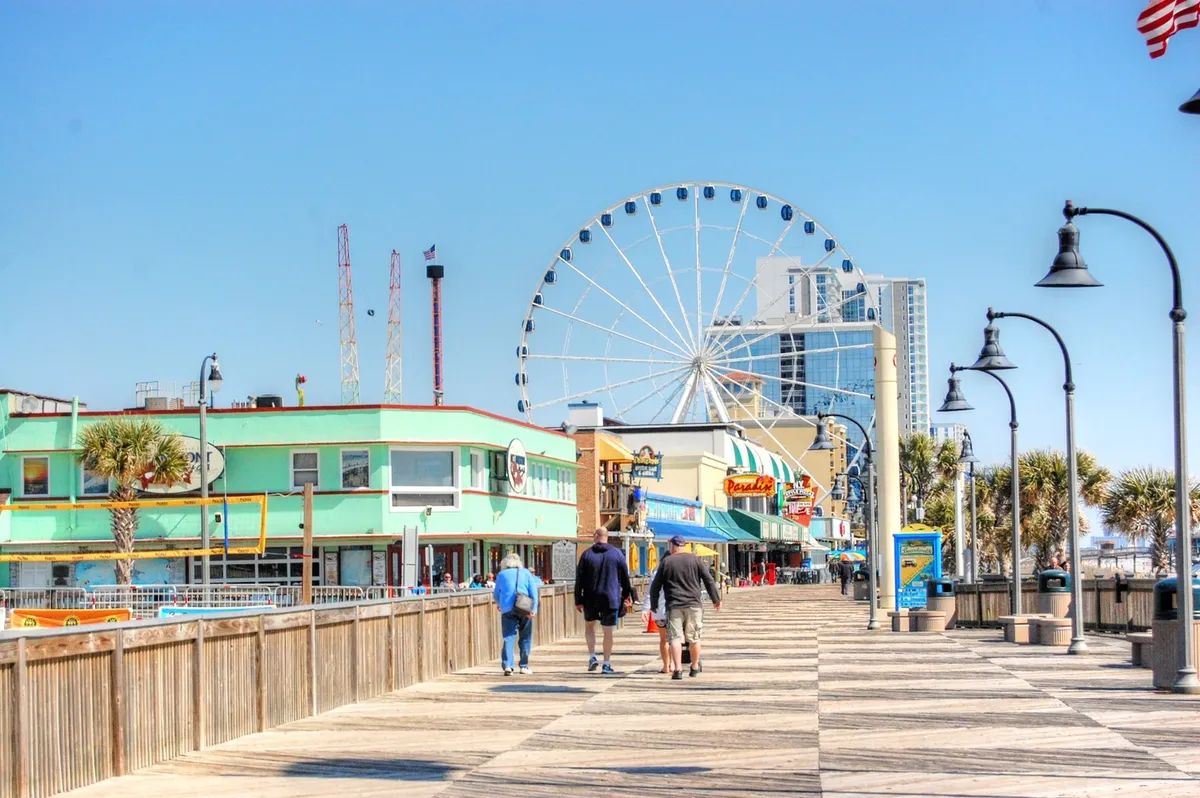 A lively boardwalk scene featuring a ferris wheel, colorful buildings, and people strolling. The area is bustling, highlighting the joy of visiting iconic Myrtle Beach spots like the SkyWheel and enjoying the vibrant atmosphere.
