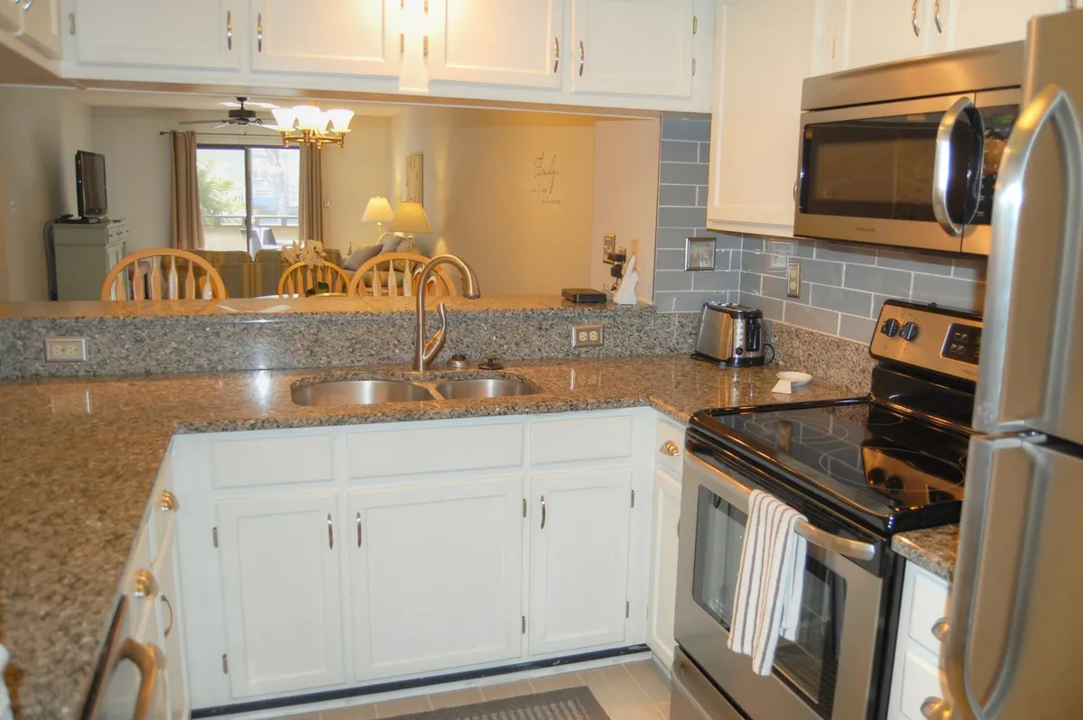 A warm kitchen scene with granite countertops, stainless steel appliances, a kitchen sink, and wooden dining chairs. The open layout suggests easy flow between cooking and dining spaces, reinforcing the idea of gathering and meal sharing.
