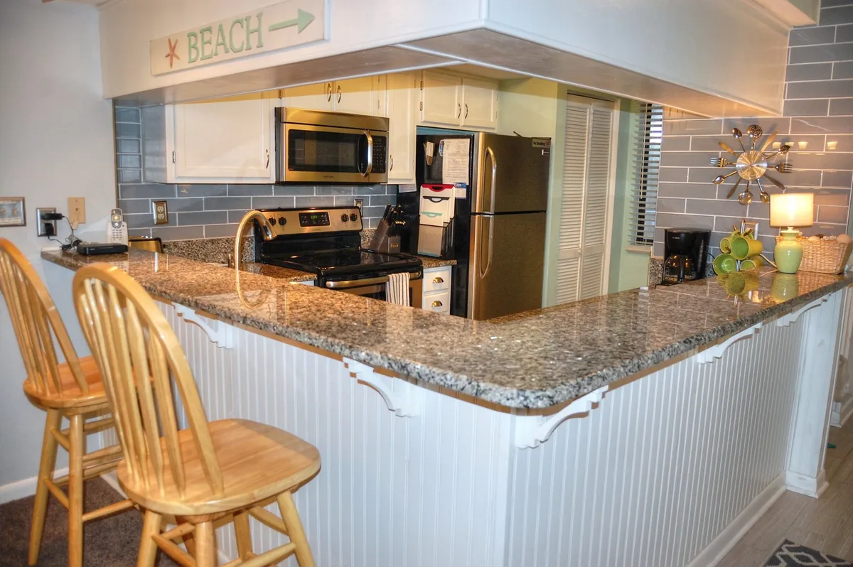 A bright, welcoming kitchen with a granite countertop, stools for casual dining, modern kitchen appliances, and decorative accents. The spacious layout encourages cooking together and socializing, with everything needed for meal prep within easy reach.