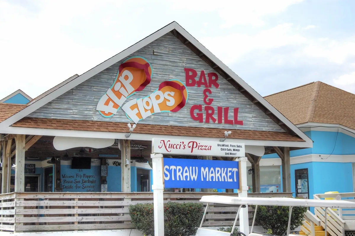 This image features the signage for Flip Flops Bar & Grill, Nucci's Pizza, and the Straw Market, set in an inviting outdoor environment. The scene is lively yet relaxed, clearly popular with locals and visitors who are looking to dine, socialize, and enjoy the coastal atmosphere.