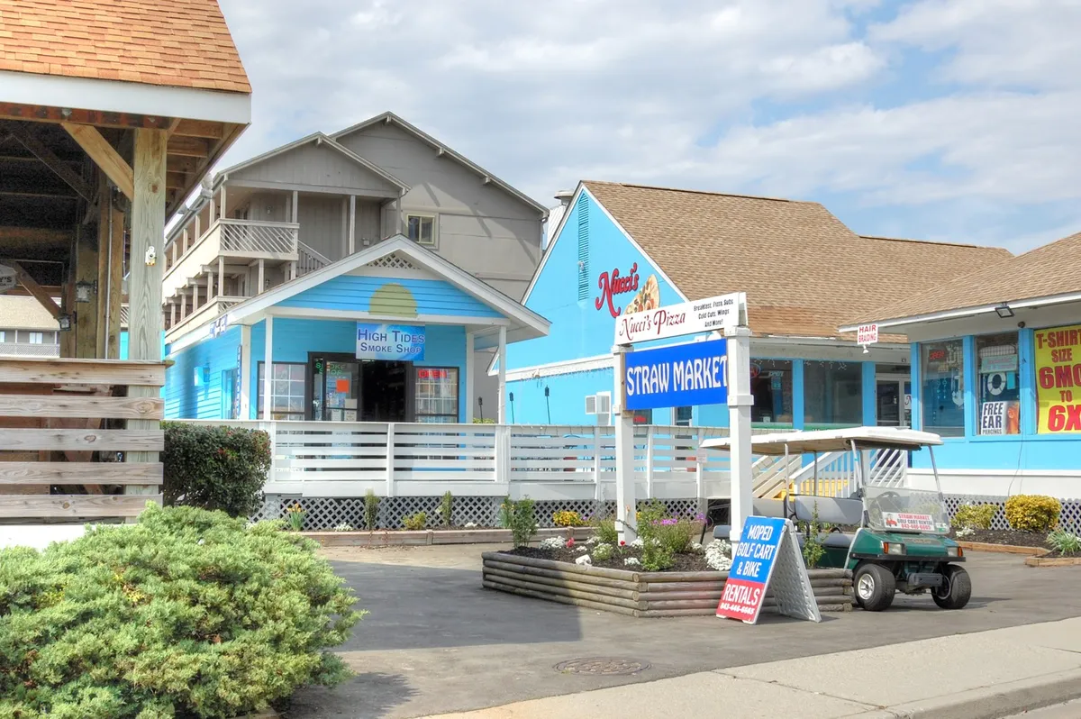 A vibrant street scene featuring colorful storefronts including Straw Market, High Tide Smoke Shop, and Nina's Pizza. People are likely browsing the shops or enjoying pizza, while the overall setting is casual and lively, perfect for capturing the local energy and opportunities for group outings.